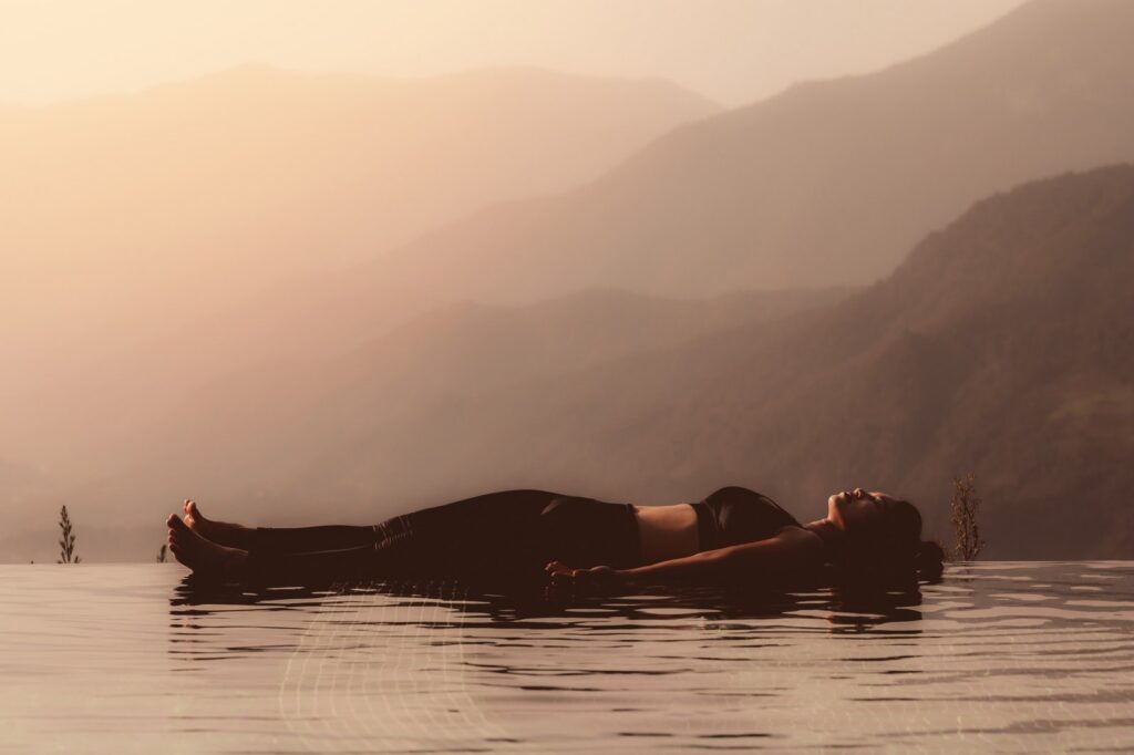 A woman lying in savasana beside a pool in morning light, with mountain views behind her.