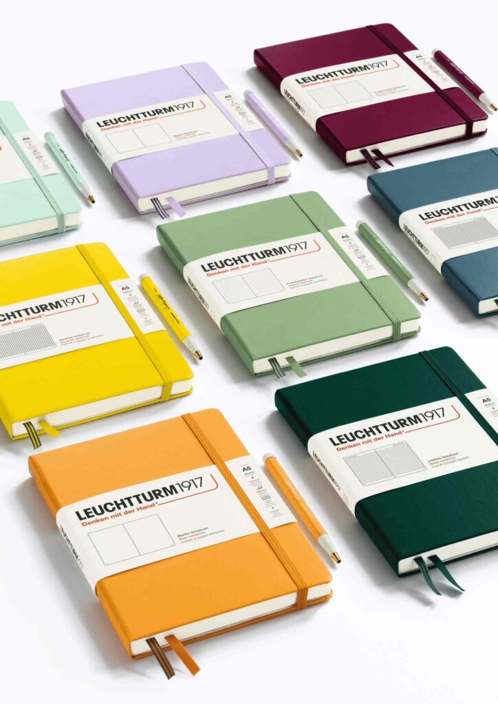 LEUCHTTURM1917 A5 hardcover notebooks in sage, lilac, port red, orange, yellow and forest green, each banded and paired with a matching pen.