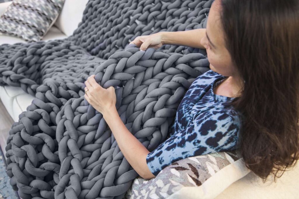 A woman under a grey knitted weighted blanket on a white couch in a bright living room.